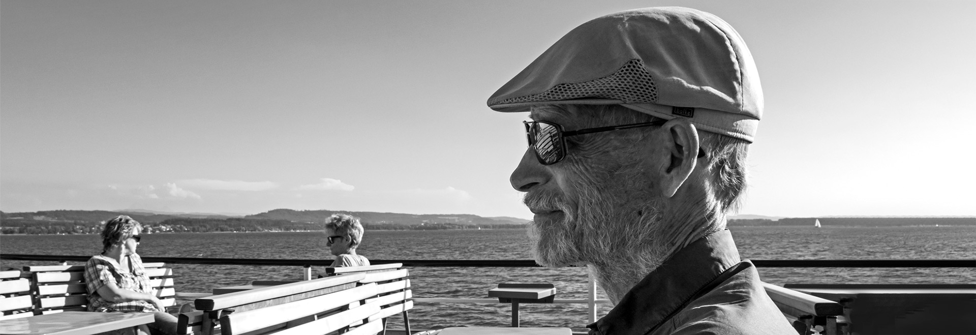 Audiodescrição (AD): Foto: Antoninho Perri. Em área externa às margens de um amplo lago, foto em preto e branco, de perfil e à curta distância de homem sentado em banco de madeira com ripas, voltado para a esquerda da imagem. À frente dele, à esquerda da foto, há 2 pessoas também sentadas em bancos, uma de frente à outra. Ao fundo de toda imagem, ampla lagoa com várias casas e áreas com vegetação em todo o contorno, e o céu quase sem nuvens em dia ensolarado. O homem usa boina e óculos escuro. Imagem 1 de 1