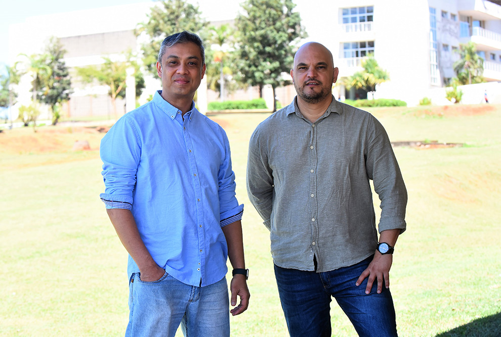 Foto de dois homens que aparecem de pé em um espaço aberto. O homem à esquerda é branco, tem o cabelo curto e está sorrindo. Ele usa calça jeans e camisa azul clara. O homem à direita é branco, careca, tem barba e bigode pretos e está usando calça jeans e camisa cinza.