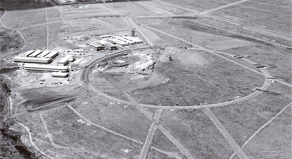 Foto em preto e branco, aérea, que mostra alguns prédios em um campo de plantação.