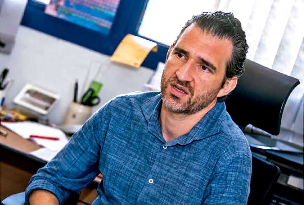 Foto de um homem sentado. Ele é branco, tem cabelo curto, barba e bigode curtos e escuros. Está usando camisa azul e tem as mãos cruzadas sobre as pernas.