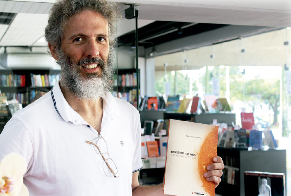 Foto de um homem de pé em um laboratório. Ele é branco, tem cabelo curto, barba e bigode grisalhos. Está usando camiseta polo branca e está segurando um livro.