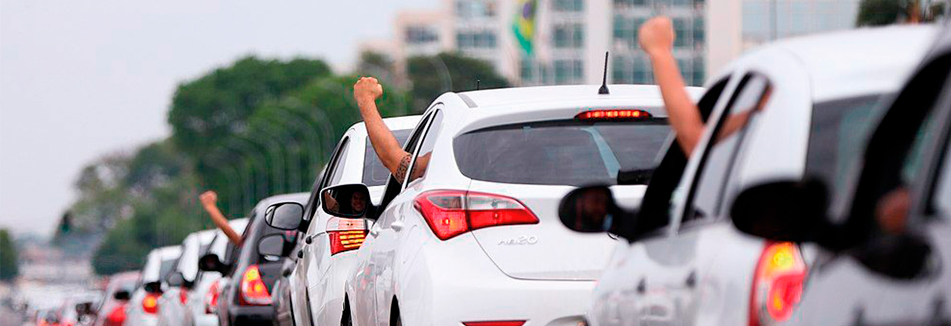 Audiodescrição. Foto: Agência Brasil/EBC. Em uma rua, imagem em perspectiva e à média distância de uma fila com 8 carros posicionados um atrás do outro, com a parte traseira em primeiro plano, seguindo da direita para o fundo da imagem. A maioria dos carros é branca e está com lanternas acesas. Três motoristas estão com o braço esquerdo para fora da janela, esticados para o alto e com punhos cerrados. Ao fundo da foto, imagem desfocada de área com vegetação, árvores e um prédio. Imagem 1 de 1