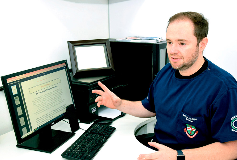 Foto de um homem em uma mesa de trabalho. Ele é branco, tem o cabelo curto e castanho, barba rala e usa camiseta azul escura