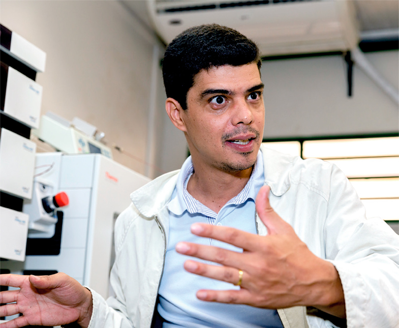 Foto de um homem que aparece do peito para cima. Ele está em um laboratório, usa avental, é branco, tem o cabelo preto e curto