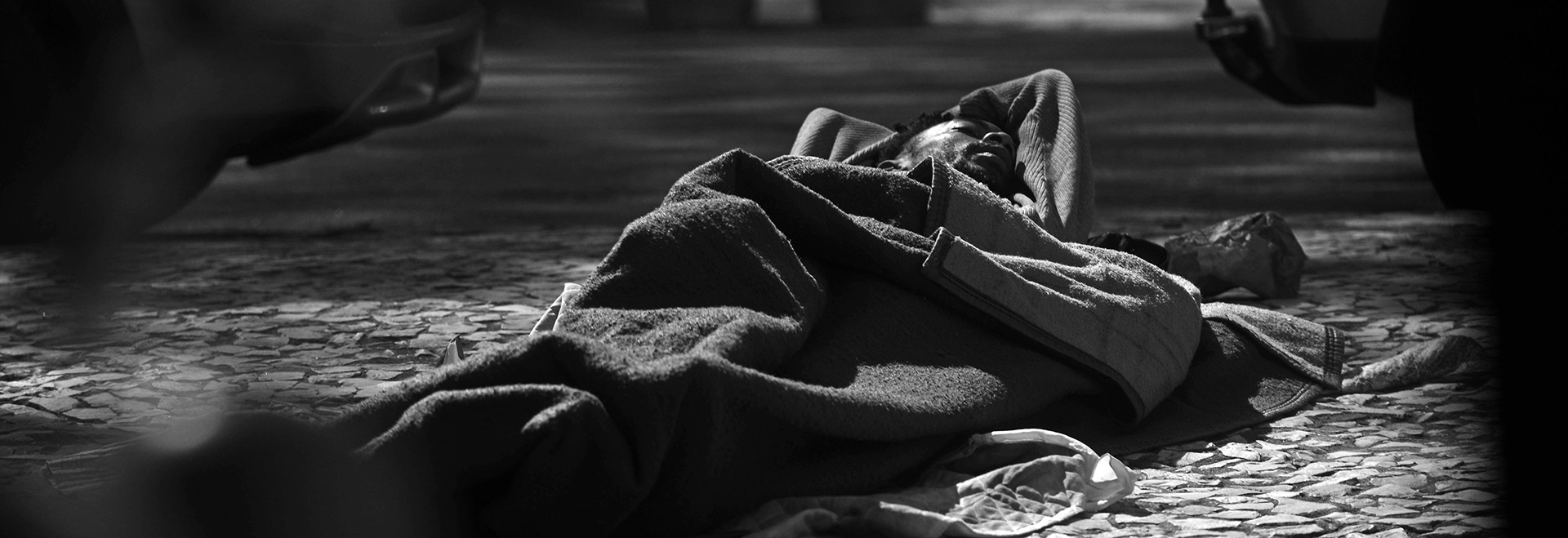 Audiodescrição. Foto: Antonio Scarpinetti. Na calçada de uma rua, imagem em preto e branco à curta distância de homem deitado no chão que dorme, virado de barriga para cima envolto em cobertores. O calçamento do local é em pedra portuguesa. Ele está com a cabeça posicionada próxima ao meio fio, na perpendicular, perto de 2 carros estacionados na rua no período noturno. Imagem 1 de 1