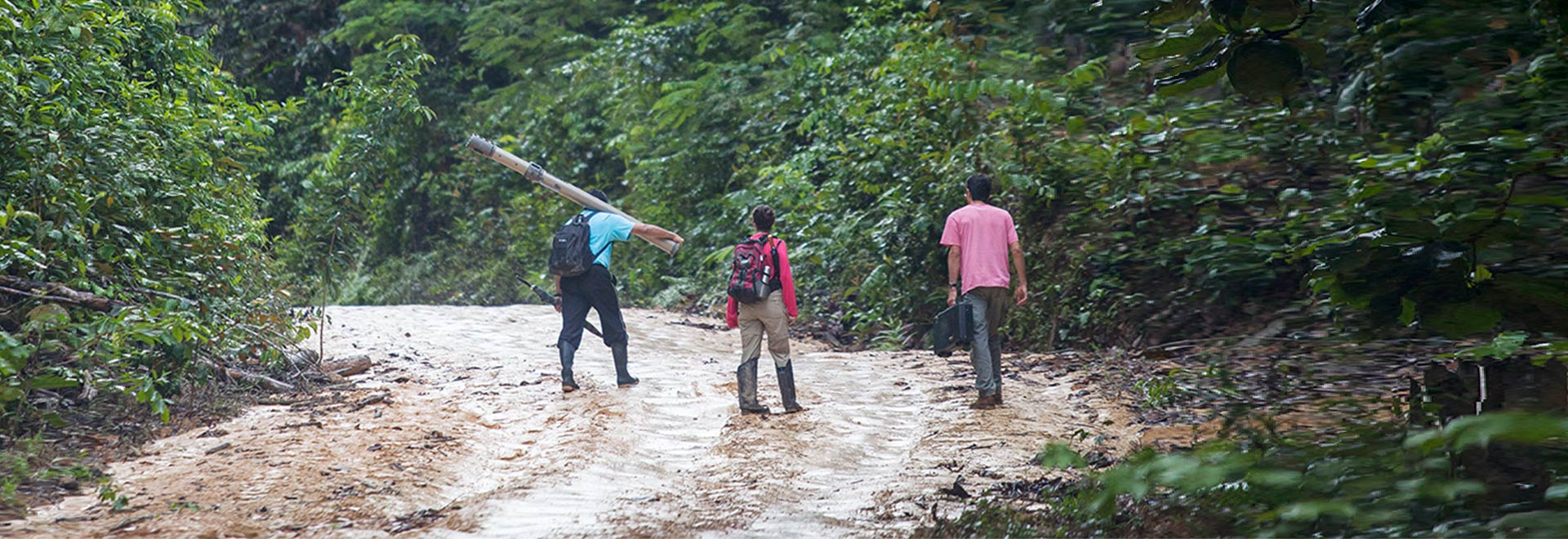 Audiodescrição. Foto: João Marcos Rosa. Em área com ampla floresta, imagem à média distância e de costas de três pessoas em pé, posicionadas uma ao lado da outra, que caminham em uma corredeira rasa de rio, com cerca de 6 metros de largura. Todos usam botas, sendo que um homem, à esquerda da foto, carrega no ombro direito equipamento semelhante a um tubo, e tem mochila às costas, assim como uma mulher, ao centro. Um homem, à direita, segura uma maleta na mão esquerda. Imagem 1 de 1