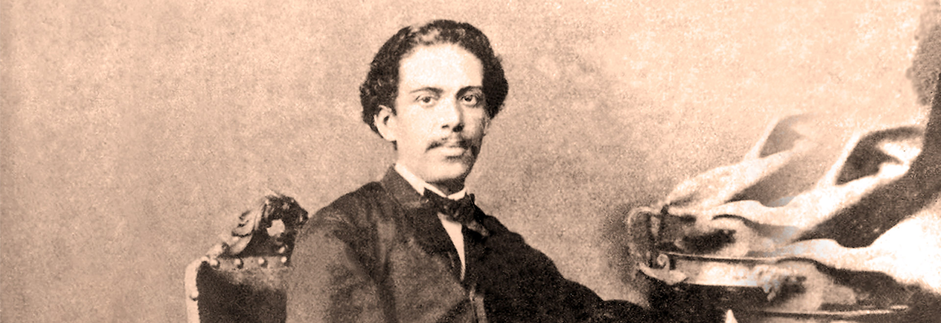 Audiodescrição. Foto: Academia Brasileira de Letras. Em uma sala, imagem centenária em tom sépia, de um homem sentado em cadeira com olhar voltado para a câmera fotográfica. A foto é à curta distância e de busto. Ele usa um tipo de gravata borboleta e terno escuros. Imagem 1 de 1