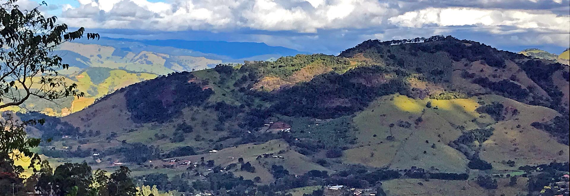 Audiodescrição. Foto: Divulgação. Imagem panorâmica de uma série de cadeias de montanhas que se estendem até o fundo da foto, recobertas por vegetação. Há algumas casas esparsas nas montanhas em primeiro plano. O céu está azul, mas também há várias nuvens. Há montanhas com áreas onde incide a luz do sol, e outras com sombra, principalmente, aquelas que aparecem em primeiro plano na foto. Imagem 1 de 1