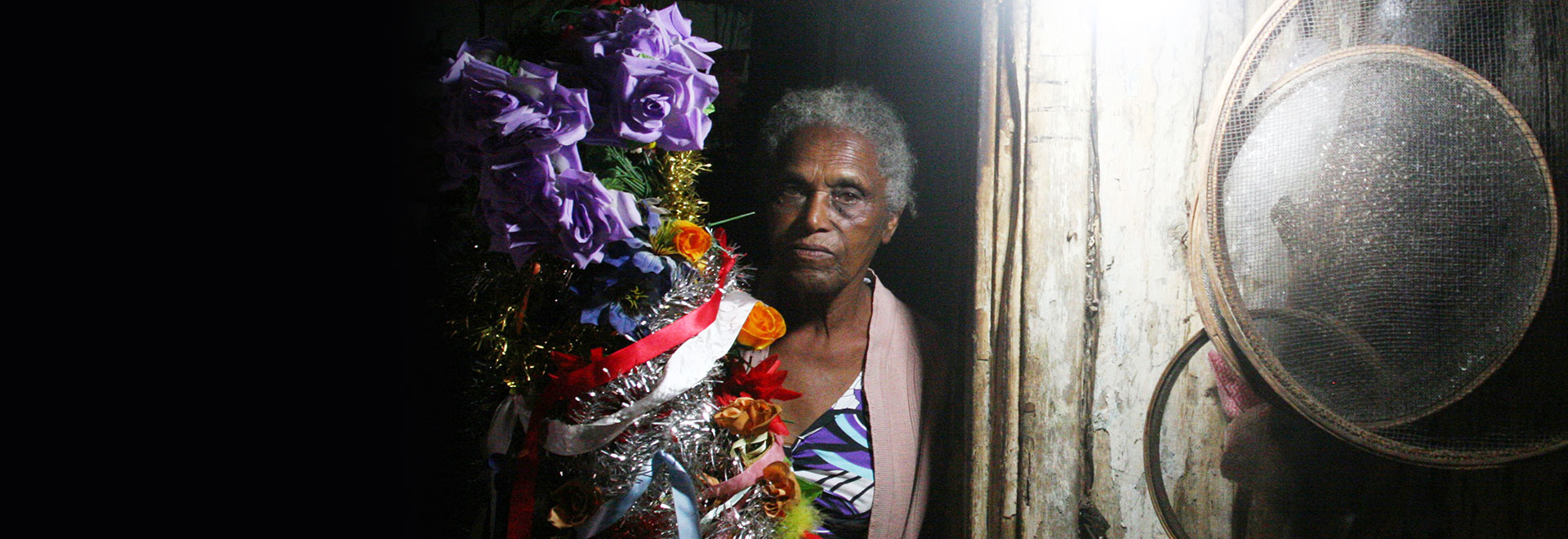 Audiodescrição. Foto: Antonio Scarpinetti. Imagem frontal e à curta distância de mulher em pé, com olhar voltado para a câmera fotográfica, que abraça junto ao lado direito do peito bandeira de Folia dos Reis formada por rosas feitas em tecido nas cores lilás, vermelho e amarelo, material semelhante a festim de natalino, nas cores prata e dourado, e fitas vermelhas e brancas. À mulher está encostada lateralmente em uma rústica porta de madeira, onde há peneiras afixadas. A imagem é noturna. Imagem 1 de 1