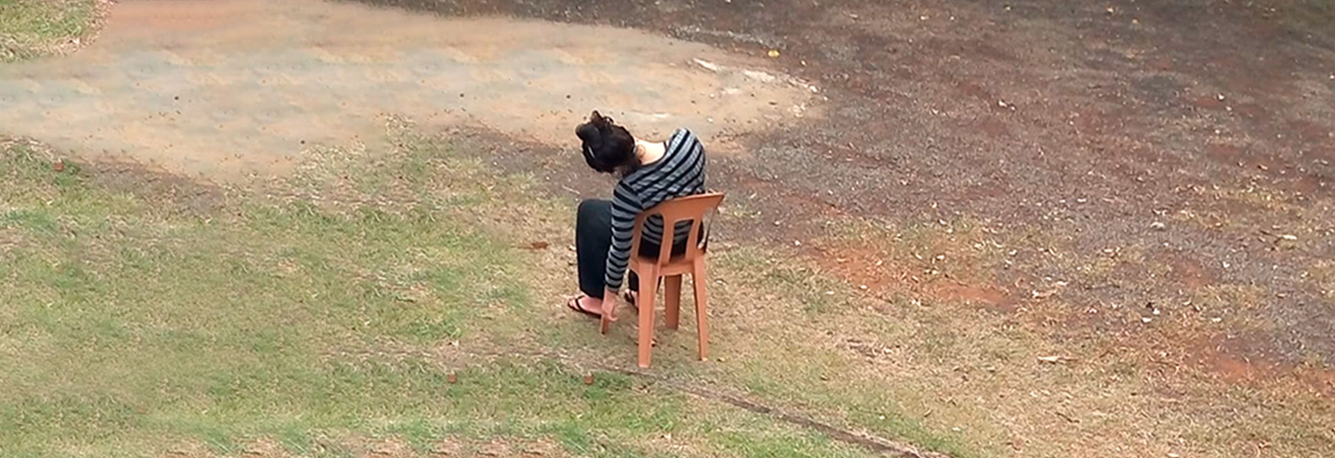 Audiodescrição. Foto: Laila Padovan. Em ampla área externa parte gramada, parte com terra, imagem à média distância, de cima para baixo, de uma pessoa sentada em uma cadeira plástica, de costas na imagem. Ela parece desmaiada, com o corpo pendendo para a esquerda da imagem, e o braço esquerdo solto, voltado para baixo, quase tocando o chão. Ela traja camisa de mangas compridas com listras horizontais em preto e cinza, calça preta e usa chinelos. Imagem 1 de 1