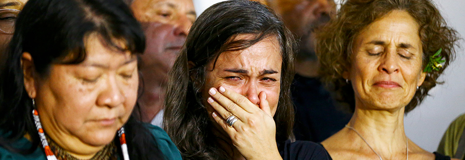 Audiodescrição. Foto: Marcelo Camargo. Em área interna, imagem frontal à curta distância de 3 mulheres em pé, posicionadas uma ao lado da outra, em foto de busto. A mulher ao centro chora profundamente, mantendo a mão esquerda no rosto, de modo a cobrir a boca, enquanto as outras mulhres estão com os olhos fechados em expressão de dor e sofrimento. Há outras pessoas em pé logo atrás delas. Imagem 1 de 1