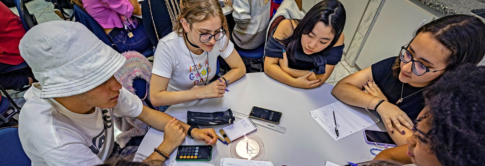 Audiodescrição. Foto: Felipe Bezerra. Em uma sala de aula, imagem a curta distância e de cima para baixo de um grupo de 4 adolescentes sentados, um ao lado do outro, ao redor de uma mesa branca redonda, com cerca de um metro e meio de diâmetro. Todos estão com olhares voltados para um objeto redondo e plástico disposto ao centro da mesa, onde também há estojos, folhas de papel, celulares e canetas. Ao fundo, outros jovens sentados ao redor de mesas. Todos usam roupas casuais. Imagem 1 de 1