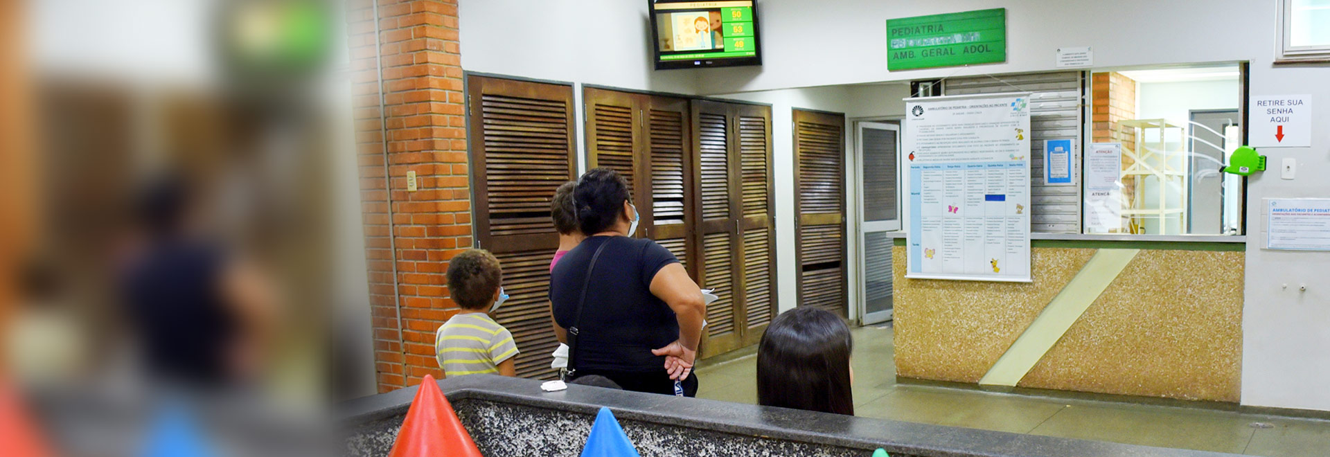 Audiodescrição. Foto: Antoninho Perri. Na recepção de um ambulatório médico, imagem a média distância e de costas de 4 pessoas em pé, voltadas para a direita da foto, sendo 3 crianças e uma mulher. À frente delas há um balcão de atendimento fechado com vidro, onde há um banner afixado, e na parte superior da parede, placa retangular verde onde se lê pediatria e uma tv lcd. À esquerda da foto, corredor de entrada ao serviço. Em primeiro plano, 4 estruturas plásticas em forma de lápis colorido gigante. 1 de 1