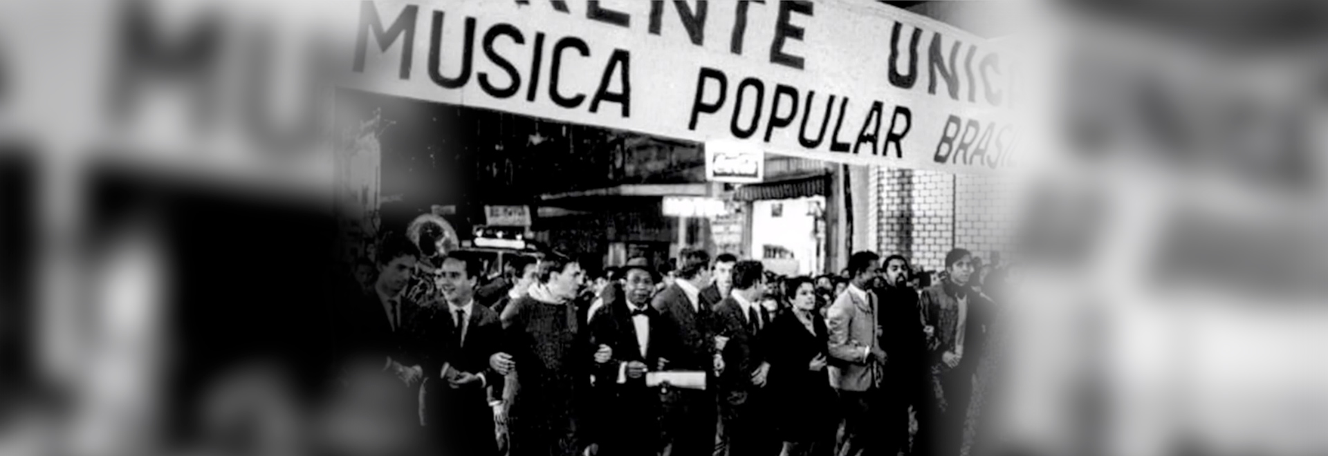 Audiodescrição. Foto: Reprodução. Imagem em perspectiva, e em preto e branco, dos anos 50/60, de grupo com cerca de 12 pessoas que caminham, da esquerda para a direita em uma rua durante a noite, sendo que estão de braços dados, uma ao lado da outra. A maioria traja terno e gravata. Há várias outras pessoas atrás delas. No alto, logo acima delas, há afixada uma extensa faixa de tecido branco onde se lê música popular brasileira. Ao fundo da foto, fachadas de estabelecimentos comerciais. Imagem 1 de 1