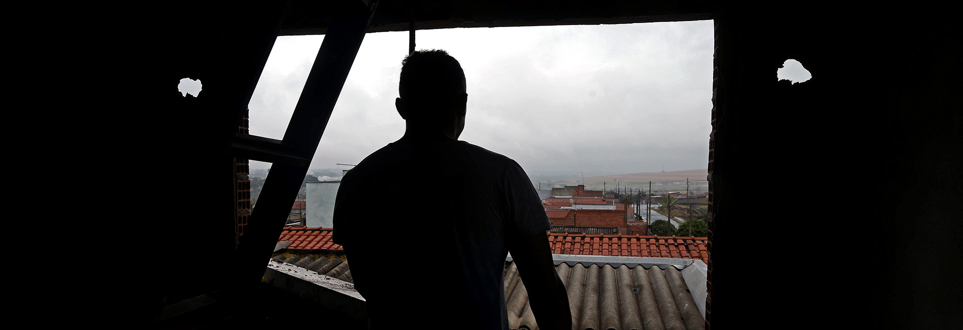 Audiodescrição. Foto: Antonio Scarpinetti. Em um casa ainda não acabada, imagem a média distância de um homem, de costas na foto, que se encontra em pé e de frente a uma janela observando a área externa, com mãos apoiadas no parapeito. Ele está no segundo andar da casa. A foto foi feita contra luz, por isso a imagem do homem e das paredes da casa são totalmente pretas. Do lado de fora, telhados de outras casas e, ao fundo, área descampada com terra. O tempo está bastante nublado. Imagem 1 de 1