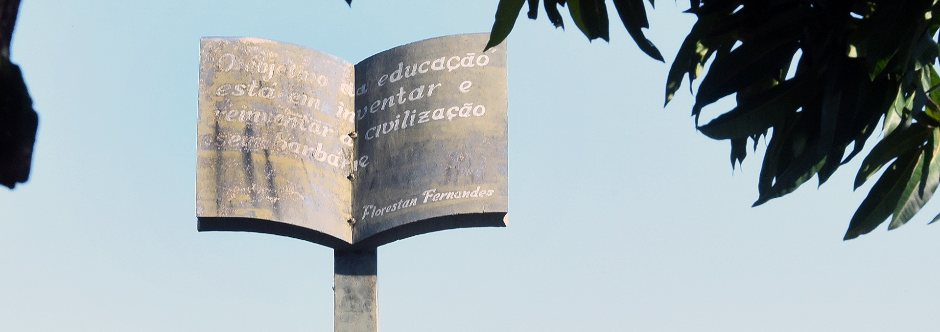 Imagem colorida: ao centro, uma escultura de livro; ao fundo, o céu e as folhas de uma árvore.