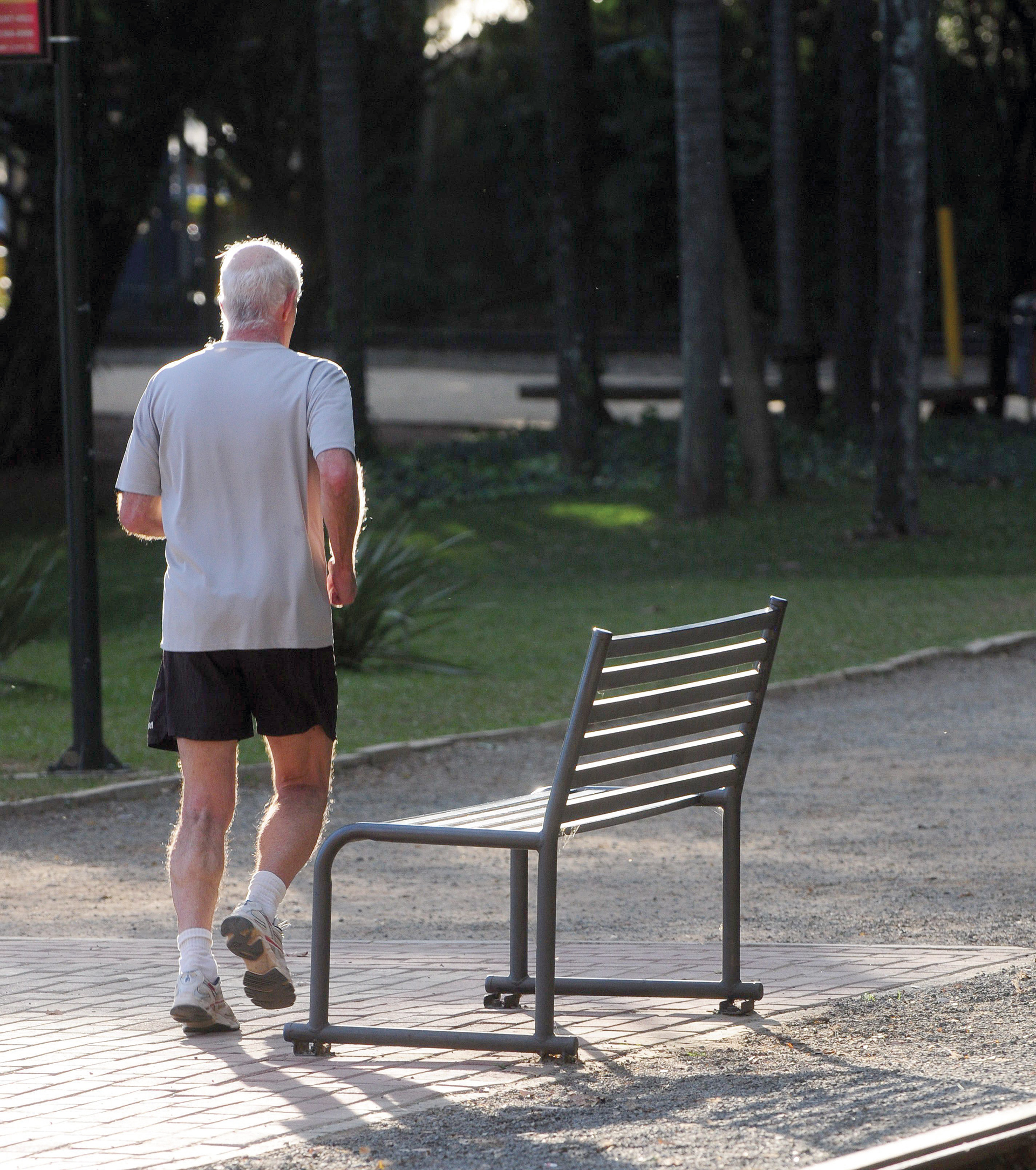 Homem idoso correndo em um parque. Ele está de costas, vestindo uma camiseta clara, shorts escuros, meias brancas e tênis. O ambiente ao redor é arborizado, com uma trilha de terra