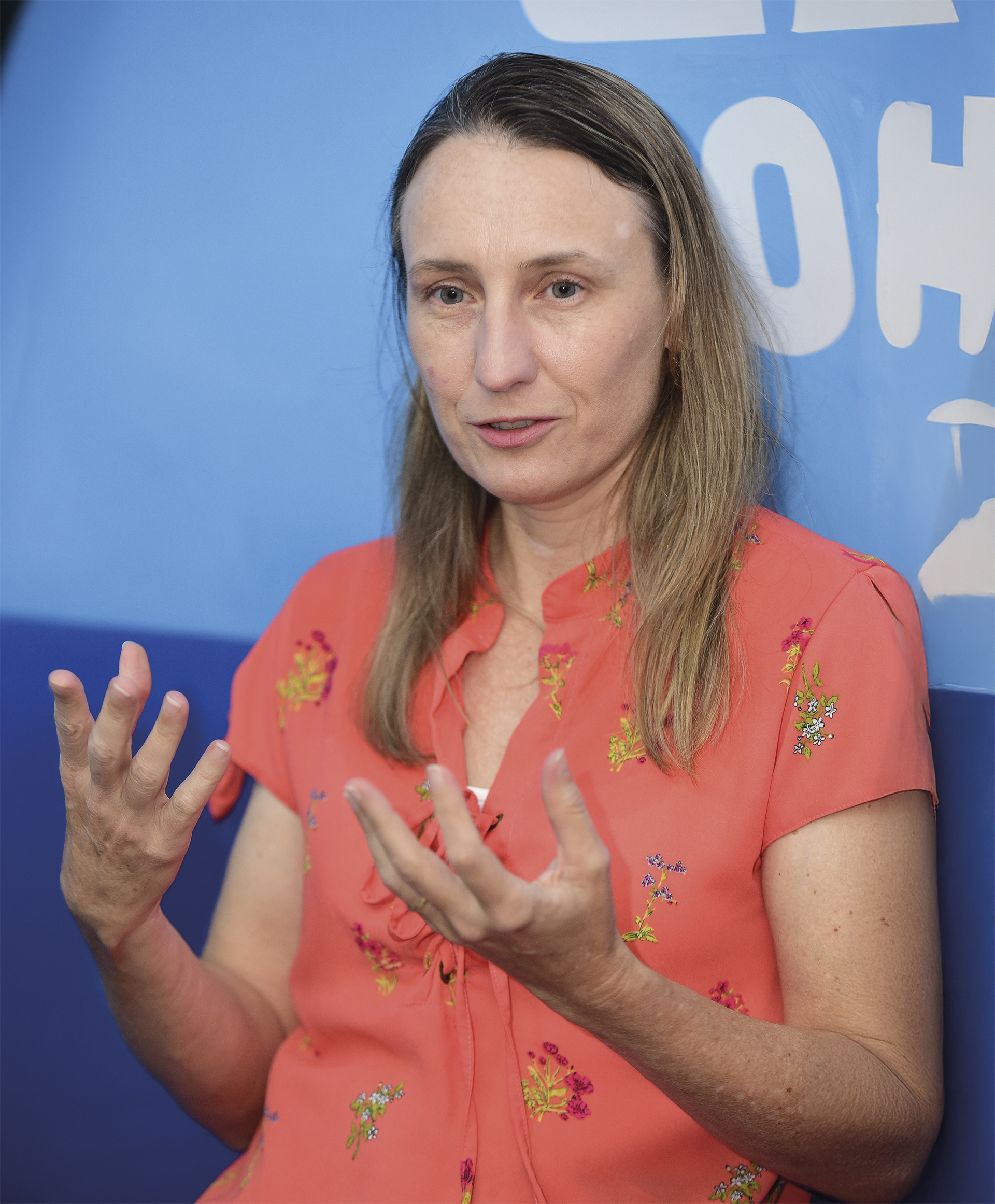Uma mulher de pele clara e cabelos lisos, castanhos claros, com mechas loiras.  A mulher está com expressão atenta e fala enquanto gesticula com as duas mãos abertas à frente do corpo, sugerindo que está explicando algo.