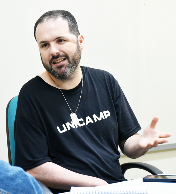 A imagem mostra um homem de barba e cabelo curto, vestindo uma camiseta preta com a palavra “UNICAMP” escrita em branco. Ele está sentado em uma cadeira, gesticulando com a mão direita enquanto fala. Ao fundo, há uma parede clara e parte de uma mesa com caderno e tablet à frente dele.