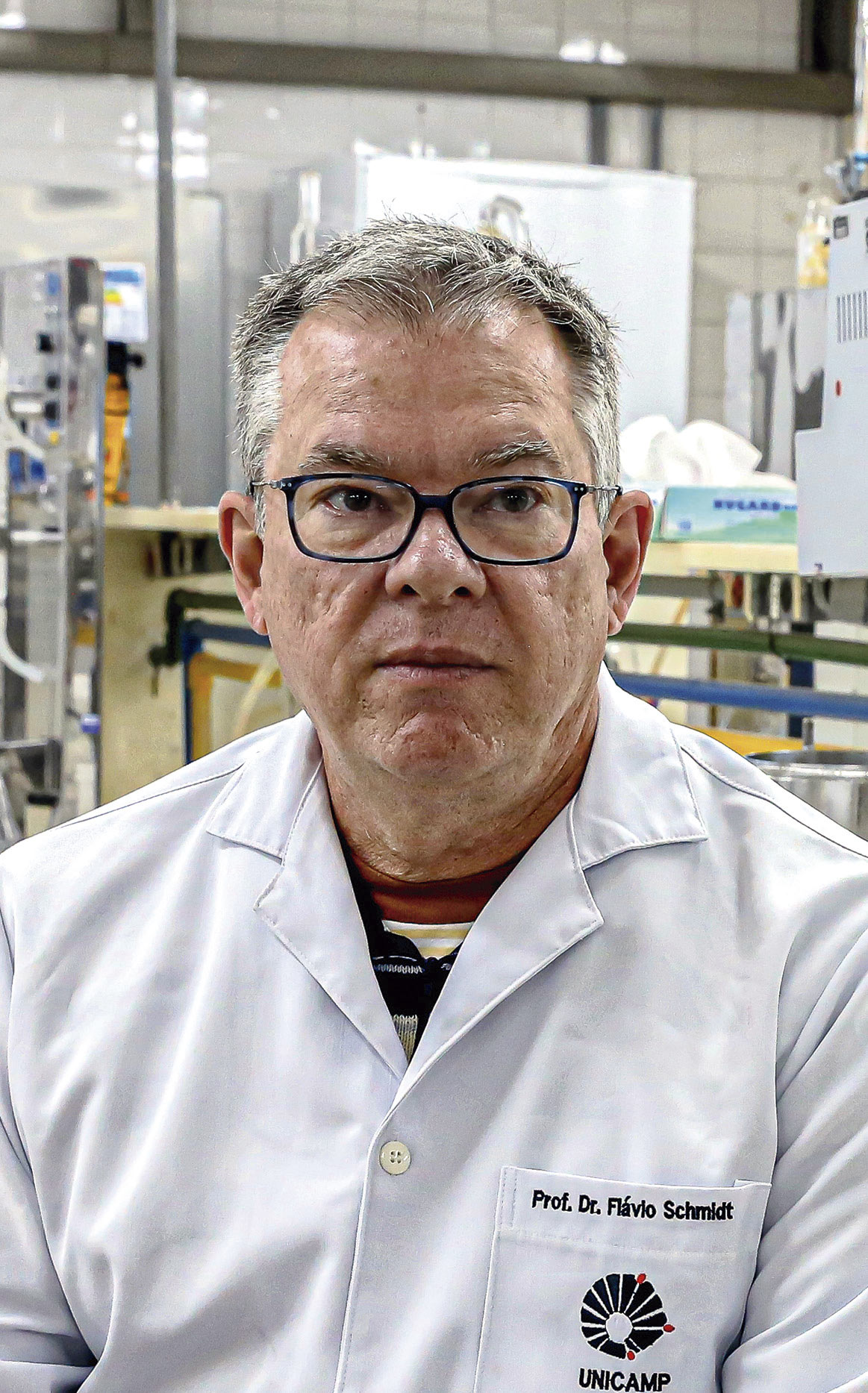A imagem mostra um homem de óculos, vestindo jaleco branco com o nome “Prof. Dr. Flávio Schmidt” e o logotipo da Unicamp, em um ambiente de laboratório.