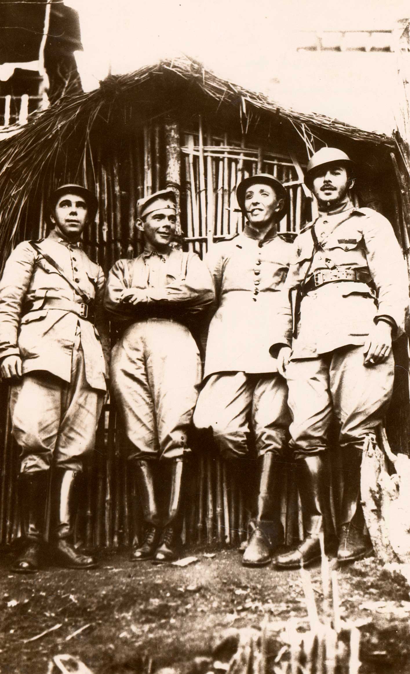 Quatro pessoas vestidas com uniformes militares posam em frente a uma construção rústica feita de bambu e palha. Os uniformes lembram estilos do início ou meados do século XX, com botas altas, cintos e capacetes. Alguns estão sorrindo, outros com os braços cruzados ou mãos na cintura, transmitindo um clima de camaradagem. Ao fundo, a estrutura parece ser uma cabana, sugerindo um cenário rural ou de época de guerra.