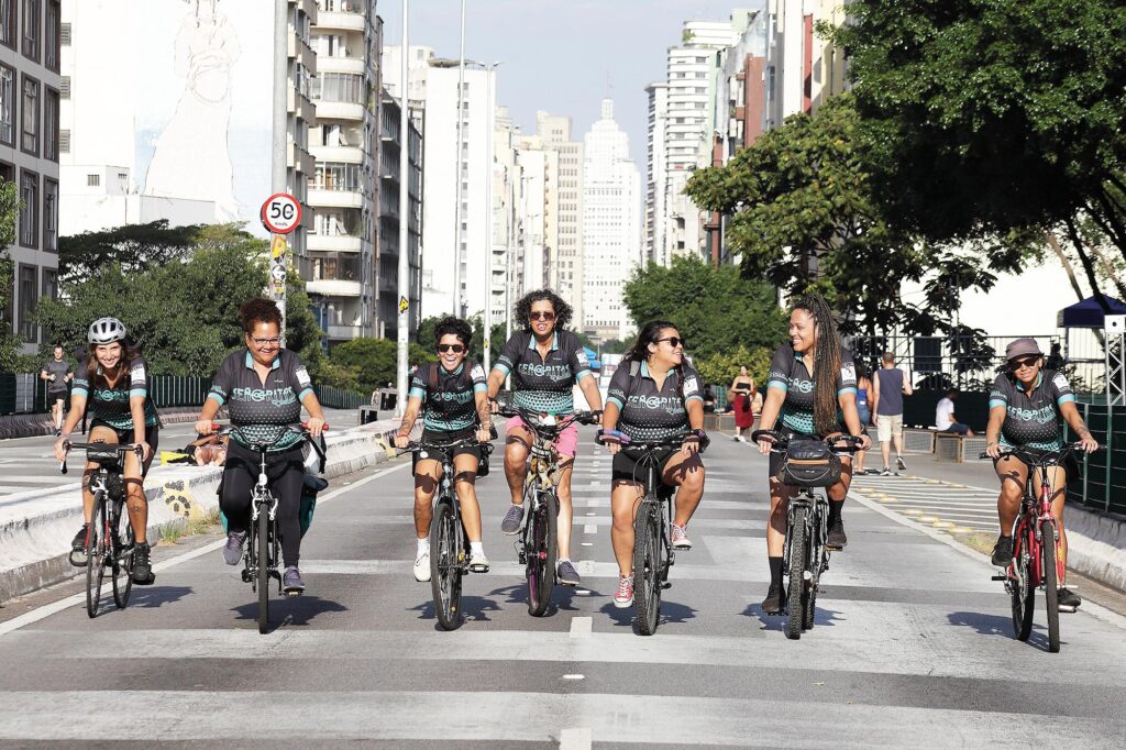 Sete mulheres andam de bicicleta em uma avenida larga, em área urbana. Todas utilizam roupas esportivas semelhantes, nas cores preto e verde. Estão lado a lado, sorrindo e interagindo umas com as outras. Ao fundo veem-se prédios altos e, mais ao fundo, o Edifício Altino Arantes (também conhecido como Banespa), ponto icônico de São Paulo.
