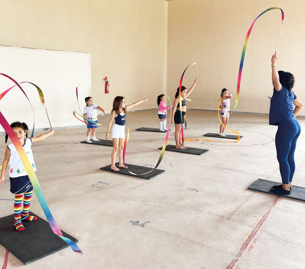 Um grupo de crianças pequenas pratica uma atividade em local fechado, acompanhadas por uma professora. Todos estão em pé sobre colchonetes pretos, segurando fitas coloridas longas, que movimentam no ar. As crianças parecem se divertir enquanto reproduzem os gestos da instrutora.