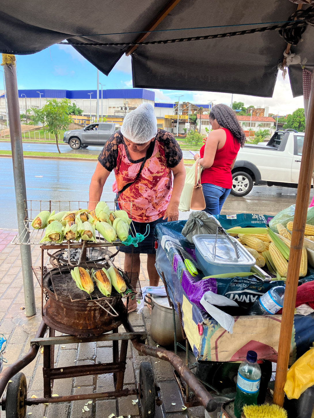 Na foto, uma vendedora de rua está sob uma cobertura, usando avental rosa e touca, enquanto assa espigas de milho em uma grelha sobre carvão. As espigas estão douradas e algumas bem tostadas. Ao lado, há uma mesa com toalha colorida, cheia de itens como garrafa de água, saco de carvão e recipientes. Outra pessoa com camisa vermelha e touca está por perto. Ao fundo, vê-se uma caminhonete branca estacionada e prédios com vegetação. O chão está molhado, sugerindo que choveu recentemente.