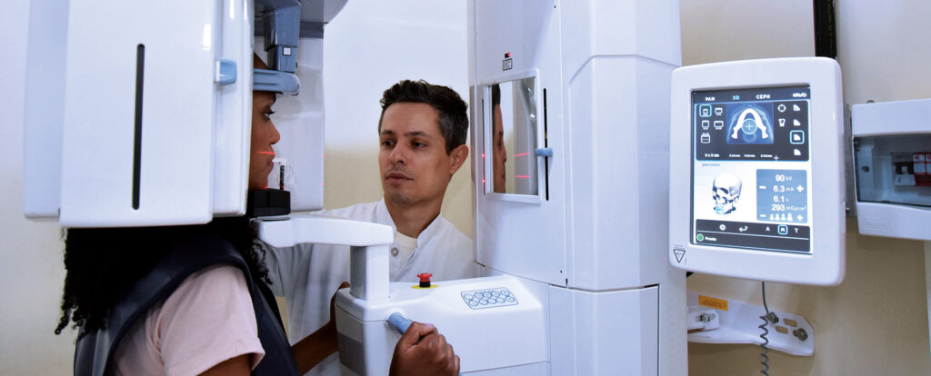 Em um ambiente clínico moderno, uum profissional de jaleco branco, identificado como Prof. Dr. Matheus de Oliveira, posiciona uma paciente em um avançado equipamento de imagem odontológica 3D, como um tomógrafo ou aparelho de raio-x panorâmico. Guias de laser vermelhas são projetadas no rosto da paciente para um alinhamento preciso, enquanto uma tela ao lado exibe a interface de controle com a imagem de um crânio e os parâmetros do exame.