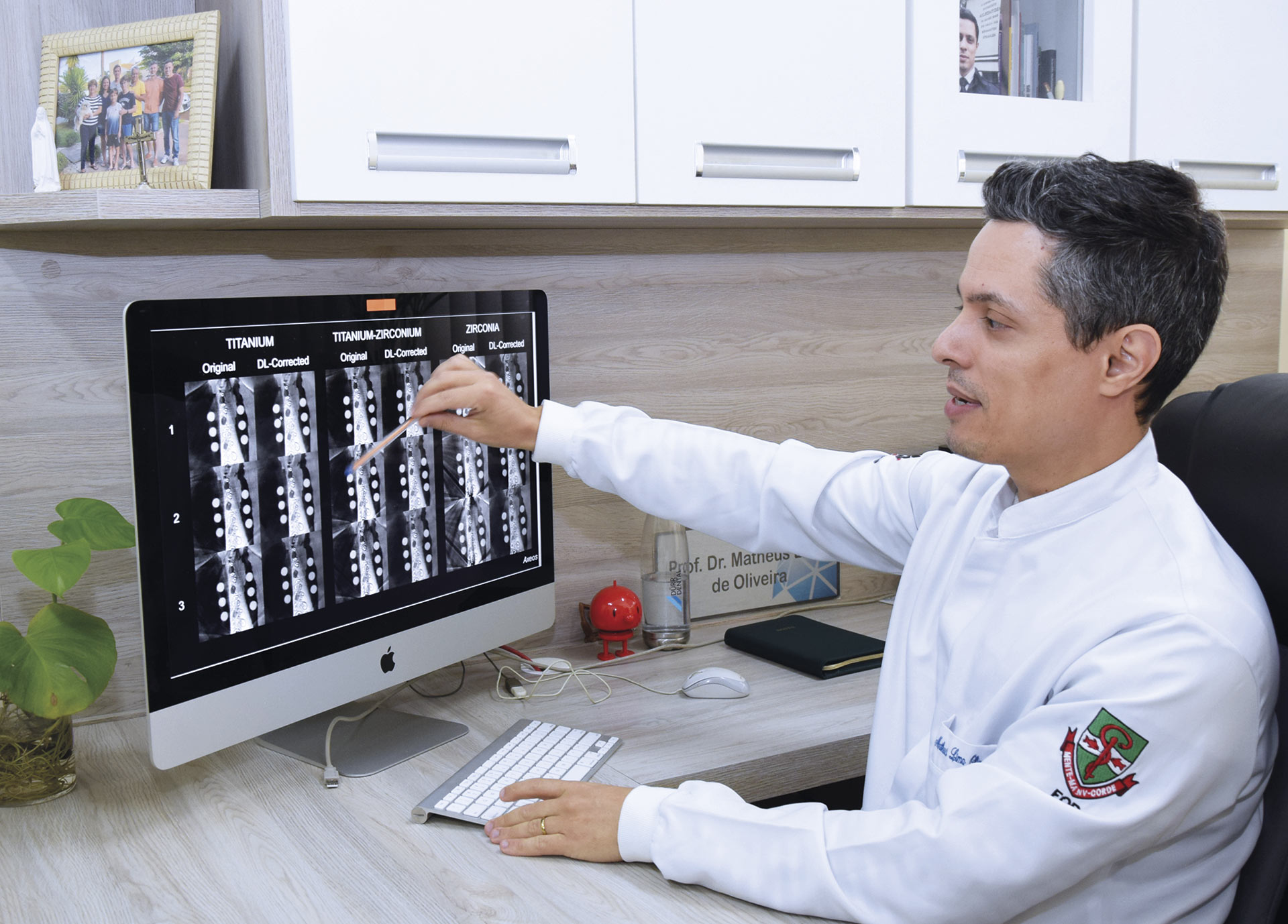Em um consultório médico ou odontológico, um profissional de jaleco branco, identificado como Prof. Dr. Matheus de Oliveira, está sentado à sua mesa e aponta com uma caneta para a tela de um computador. No monitor, são exibidas várias imagens de exames, como radiografias ou tomografias, que parecem comparar diferentes tipos de implantes dentários (titânio e zircônia). O ambiente é organizado e profissional, com armários brancos e objetos pessoais, como um porta-retrato de família, ao fundo.