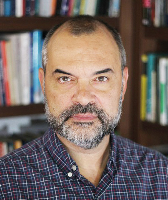 A imagem mostra um homem de pele clara, barba e cabelo curto grisalho, vestindo camisa xadrez escura. Ele está em um ambiente interno, com estante de livros desfocada ao fundo, olhando diretamente para a câmera com expressão neutra