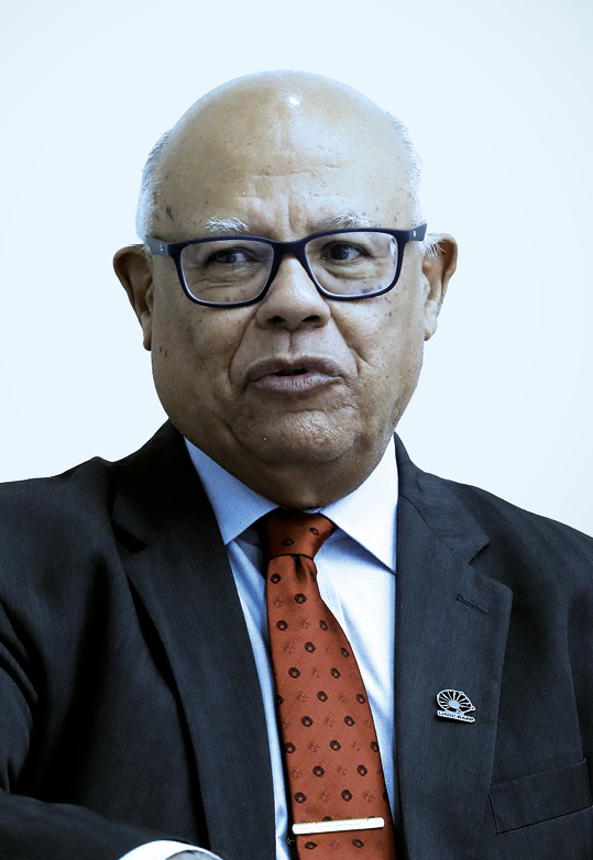 Homem usando óculos de grau e terno escuro. Ele está com uma gravata vermelha estampada e ao paletó há um pin metálico, ele está olhando levemente para o lado