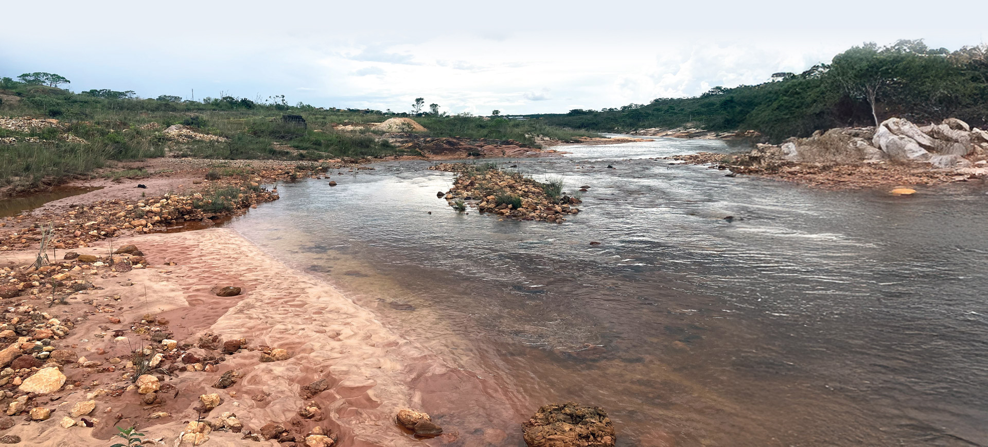 Paisagem natural mostrando um leito de rio raso e de águas escuras, que corre entre margens arenosas e pedregosas. A areia tem tonalidade avermelhada, com muitas pedras espalhadas. Em volta do rio há vegetação baixa e árvores esparsas, sob céu claro com algumas nuvens.