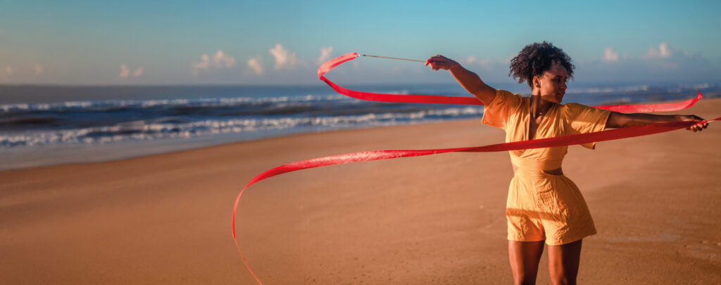 Uma pessoa está em pé na areia da praia, perto do mar, fazendo uma apresentação com uma fita vermelha longa. Ela veste um macacão curto laranja claro e está em movimento, com a fita formando curvas no ar. O céu está azul com algumas nuvens, e o mar tem ondas suaves ao fundo. A cena transmite leveza e liberdade.