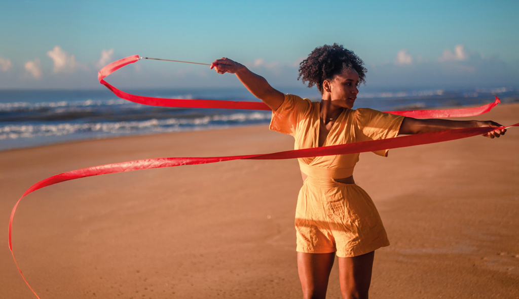 Uma pessoa está em pé na areia da praia, perto do mar, fazendo uma apresentação com uma fita vermelha longa. Ela veste um macacão curto laranja claro e está em movimento, com a fita formando curvas no ar. O céu está azul com algumas nuvens, e o mar tem ondas suaves ao fundo. A cena transmite leveza e liberdade.