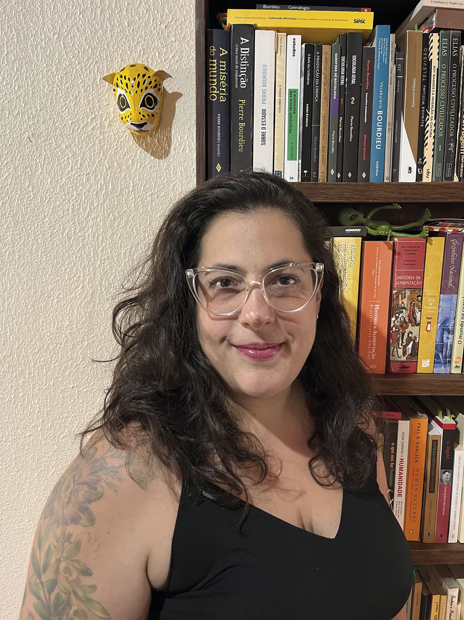 Retrato de uma mulher de cabelos escuros e óculos, com uma tatuagem no braço, em frente a uma estante de livros.