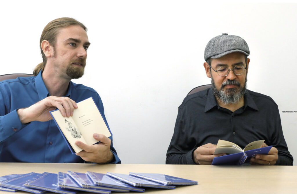 A foto mostra dois homens sentados lado a lado em uma mesa clara, ambos manuseando livros de capa azul.
À esquerda, um homem de barba curta e cabelo preso em rabo de cavalo, vestindo uma camisa azul, segura um livro aberto e parece estar mostrando seu conteúdo.
À direita, um homem de barba grisalha, óculos redondos e boina cinza lê atentamente outro exemplar. Ele veste uma camisa preta.
Sobre a mesa, vários livros iguais estão empilhados em leque, sugerindo o lançamento ou a apresentação de uma obra. O fundo é branco e simples, destacando os dois personagens e os livros.