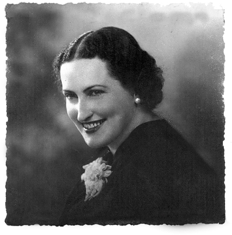 A fotografia em preto e branco mostra uma mulher em pose de estúdio, com ar elegante e sorridente.
Ela tem cabelos escuros, penteados para trás e presos, usa brincos de pérola e veste uma roupa escura adornada com um broche ou flor clara na altura do peito. O fundo é neutro e desfocado, destacando a figura.
A imagem tem as bordas levemente irregulares, características de fotografias antigas, transmitindo um ar de retrato clássico e atemporal.
