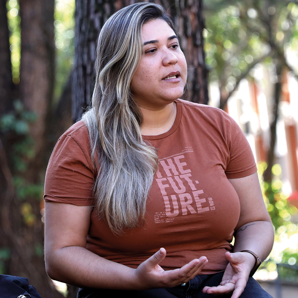 Mulher de pele clara, cabelos longos e lisos com mechas loiras, veste uma camiseta marrom com a frase “THE FUTURE” estampada em letras grandes e claras. Ela está sentada ao ar livre, com fundo de árvores, e faz um gesto com as mãos, como se estivesse explicando ou conversando sobre algo. Sua expressão é séria e atenta.