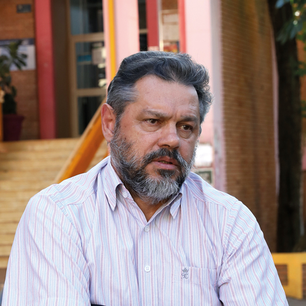 Homem de meia-idade, cabelos curtos e barba grisalha, veste uma camisa listrada em branco e rosa com pequenos detalhes. Ele está sentado ao ar livre, em frente a uma construção colorida com paredes vermelhas e amarelas e degraus de escada. Seu semblante é sério e reflexivo, olhando ligeiramente para o lado.