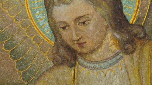 A imagem mostra um detalhe de uma pintura sacra. Nela, vemos o rosto sereno de um anjo, com cabelos longos e ondulados, de tom castanho claro. O anjo possui uma auréola dourada com fundo azul e raios decorativos em volta da cabeça, além de asas estilizadas com penas delineadas em dourado. A expressão do anjo é suave e contemplativa, transmitindo calma e espiritualidade. O fundo da pintura tem tons terrosos, destacando a figura central.