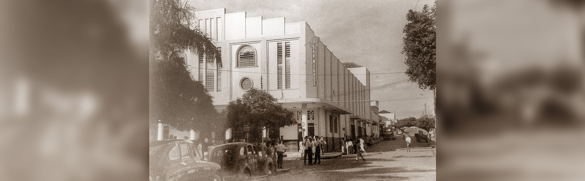 A imagem em tom sépia mostra a fachada de um prédio de arquitetura antiga, identificado como “Cine São Miguel” em uma placa vertical na esquina. Em frente, há várias pessoas reunidas na calçada e carros antigos estacionados na rua. Árvores aparecem nas laterais, e o cenário sugere uma cena urbana de meados do século XX