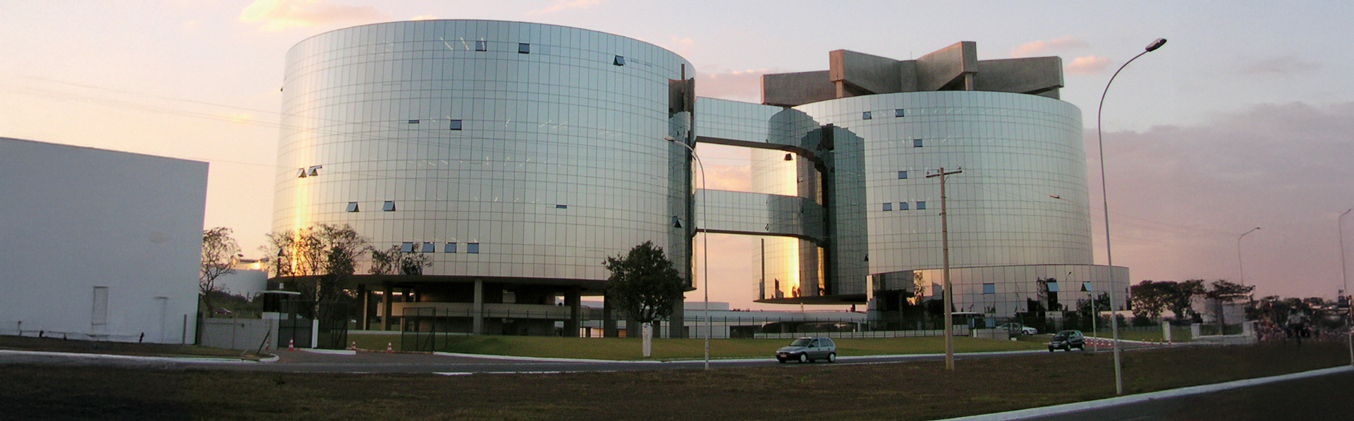 A imagem mostra um grande edifício moderno formado por duas torres cilíndricas de vidro espelhado, conectadas por passarelas elevadas. A fachada reflete o céu no fim de tarde, com tons dourados e rosados. O prédio está cercado por área verde e vias de acesso, com alguns carros estacionados e postes de iluminação ao redor.