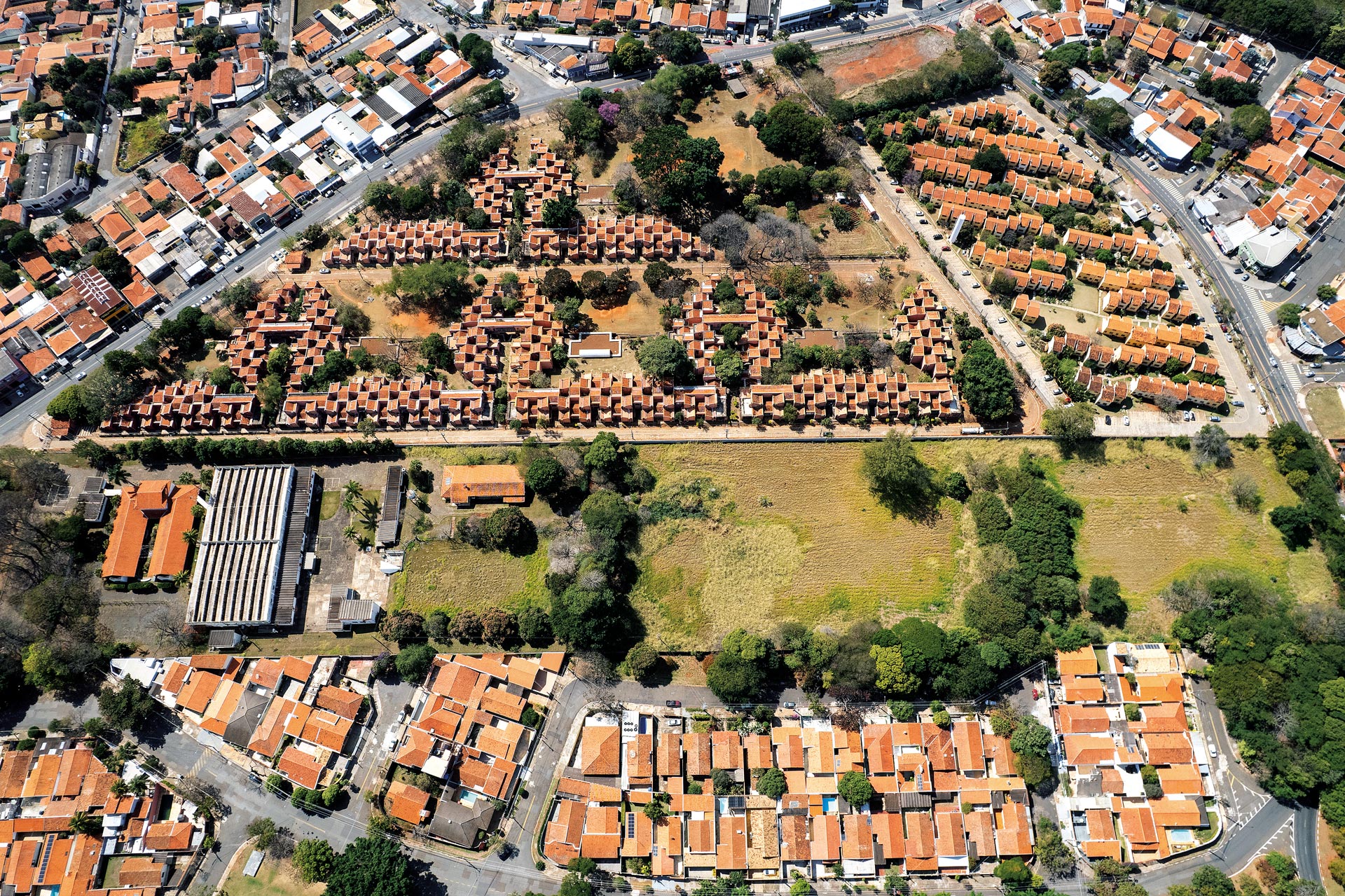 Vista aérea de um bairro com muitas casas de telhados vermelhos. Ao centro, há um grande conjunto residencial organizado em fileiras geométricas, rodeado por árvores e áreas verdes. Algumas quadras vizinhas também aparecem na imagem.