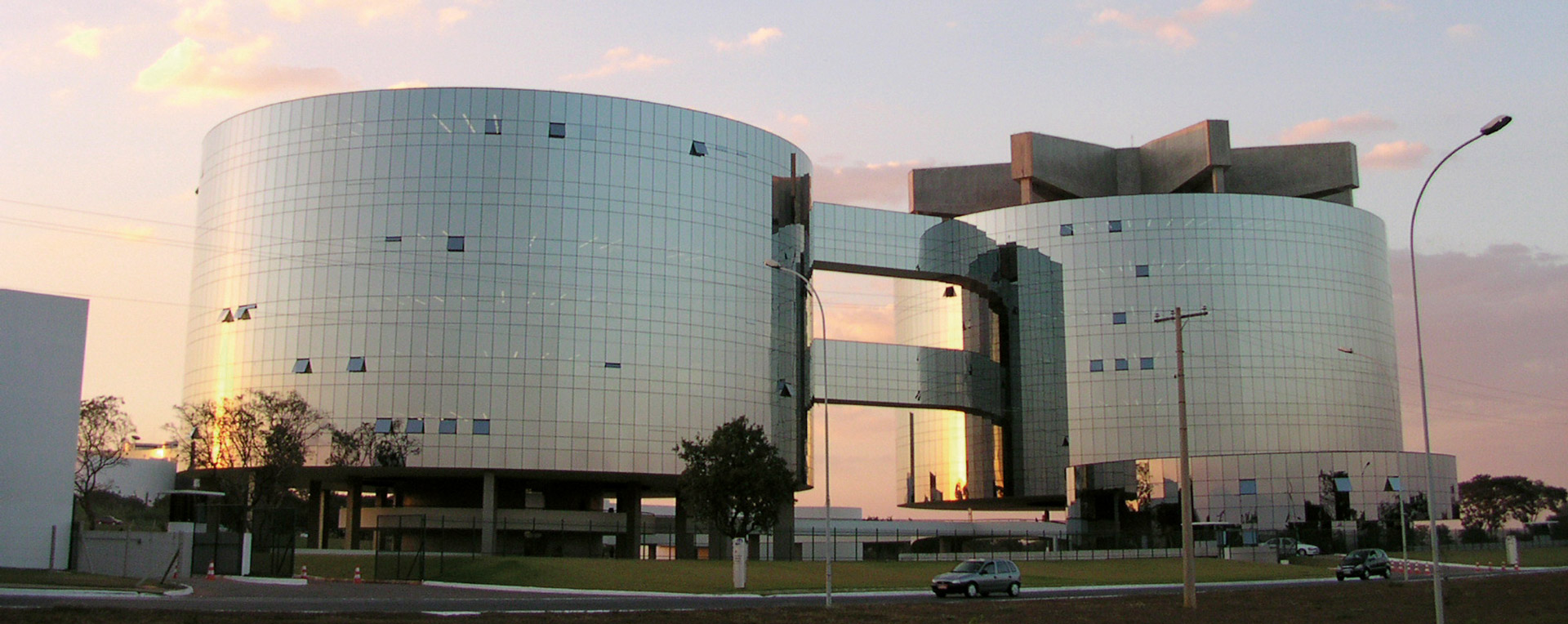 A imagem mostra um grande edifício moderno formado por duas torres cilíndricas de vidro espelhado, conectadas por passarelas elevadas. A fachada reflete o céu no fim de tarde, com tons dourados e rosados. O prédio está cercado por área verde e vias de acesso, com alguns carros estacionados e postes de iluminação ao redor