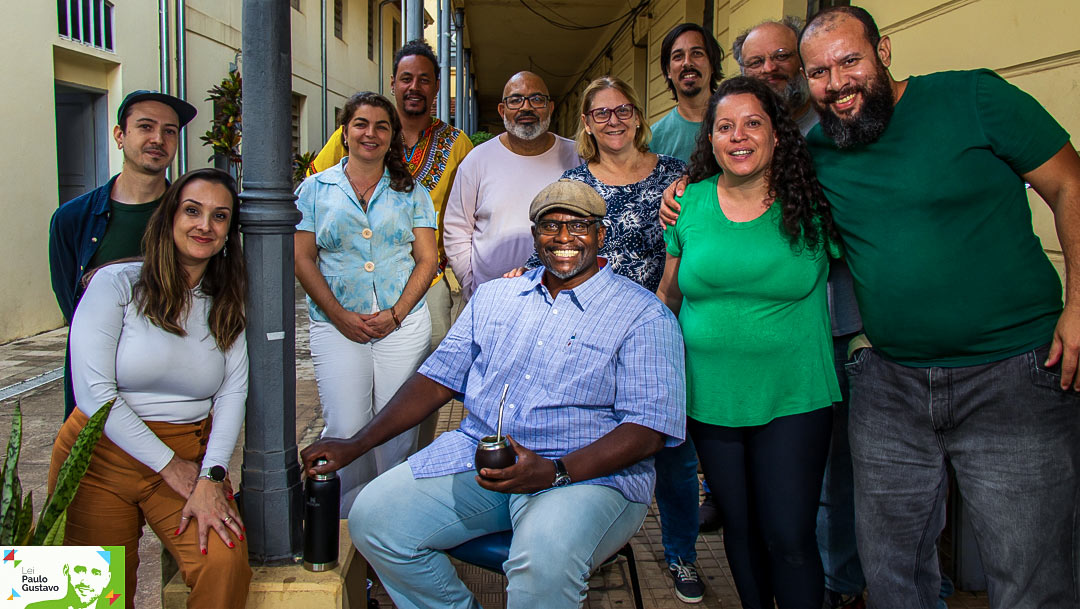 A fotografia colorida mostra um grupo de 12 pessoas reunidas em uma passagem entre prédios, sorrindo para a câmera. No centro, em primeiro plano, um homem negro, de boné bege e camisa azul xadrez, está sentado, sorridente, segurando uma cuia de chimarrão. Ao lado dele, de pé e próximos, estão homens e mulheres de diferentes idades, com roupas casuais e expressões alegres.
À esquerda, uma mulher de blusa branca e calça marrom se apoia em um poste; atrás dela e ao redor, o grupo se organiza de forma descontraída. No canto inferior esquerdo aparece o logotipo da Lei Paulo Gustavo, indicando que a foto está ligada a um projeto cultural financiado por essa lei.