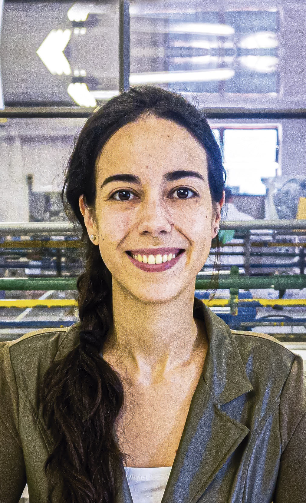 A imagem mostra uma mulher jovem sorrindo, com cabelo escuro preso em trança lateral, usando blazer verde-oliva, em um ambiente interno com equipamentos e estruturas metálicas ao fundo.