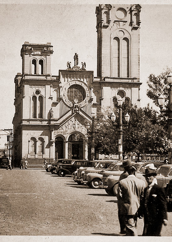 A fotografia em preto e branco mostra a fachada da Igreja Nossa Senhora do Rosário, em Campinas. Ela tem duas torres frontais — a da direita é alta e imponente, enquanto a da esquerda está incompleta e mais baixa. No centro da fachada há uma grande janela redonda, em forma de rosácea, acima da porta principal. O estilo é clássico e ornamentado, com esculturas religiosas no topo. Em frente à igreja, na rua, há alguns carros antigos estacionados e algumas pessoas andando, vestidas com roupas elegantes, como ternos e chapéus. Também há postes de iluminação ao longo da calçada. A cena transmite a atmosfera de uma cidade de meados do século 20.