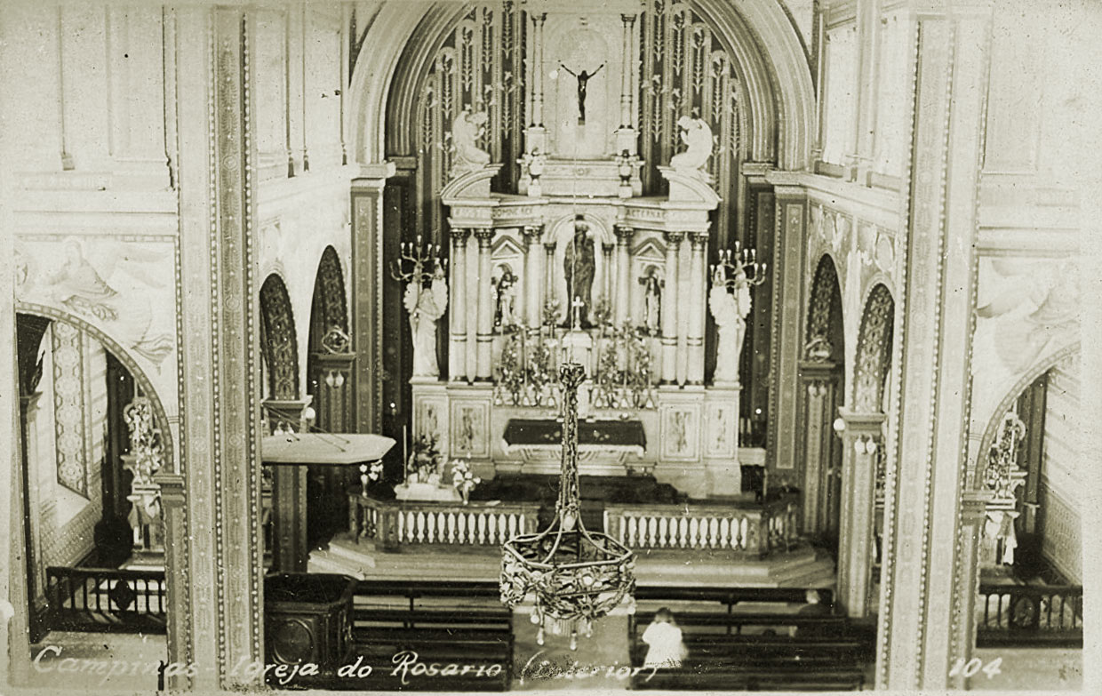 O ChatGPT disse:
A fotografia em preto e branco mostra o interior da Igreja Nossa Senhora do Rosário, em Campinas. No centro da imagem está o altar-mor, ricamente ornamentado. Ele possui colunas, nichos com imagens de santos e, no topo, um crucifixo em destaque. Há também esculturas de anjos ajoelhados e grandes candelabros de vários braços ladeando o altar. Um lustre de metal trabalhado e pendurado no teto aparece em primeiro plano. As paredes e arcos ao redor são decorados com relevos e pinturas, reforçando o caráter solene e sagrado do espaço. Nos bancos de madeira, à frente do altar, é possível ver algumas pessoas sentadas ou em movimento.