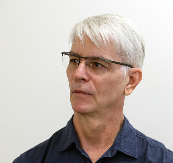 Um homem com cabelo branco e óculos de armação fina, olha para o lado esquerdo com uma expressão séria. Ele usa uma camisa azul escura. O fundo é uma parede branca.